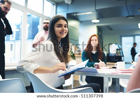 Young female manager writes important point of reference in time to the project meeting with colleagues. Attractive girl trainee keeps records for mastering new skills from the experts during briefing