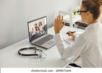 Young Female Mecical Worker In White Uniform Greeting Happy Family With Child Before Online Consultation On Laptop From Medical Clinic Office. Telehealth, Telemedicine Cocnept