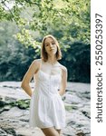 A young female model elegantly poses in a flowing white dress surrounded by peaceful and serene natural beauty