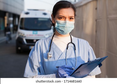 Young female NKS UK EMS doctor in front of healthcare ICU facility,wearing protective face mask holding medical patient health check form,Coronavirus COVID-19 pandemic outbreak crisis PPE shortage 