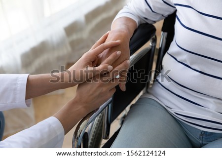 Young female nurse caregiver hold hand support disabled handicapped senior adult grandma patient sit on wheelchair, old paralyzed people with disability medical help assistance concept, close up view