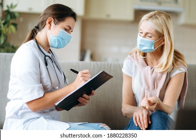 Young Female Nurse In A Medical Mask Help Middle Aged Woman During During Illness, Female Doctor Talk With Senior Patient Give Empathy Support And Prescribes Treatment While Sitting On Sofa At Home