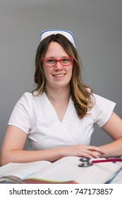 Young  Female Nurse Portrait Wearing White Scrubs, Cap With Nursing Textbooks On The Table, Smiling