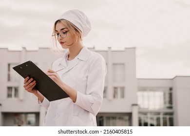Young female nurse standing outside hospital infirmary writing patient illness history in journal. Doctor woman dressed white medical gown and cap wear eyeglasses posing, fill patient paper document