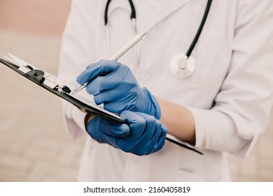 Young female nurse standing outside hospital infirmary writing patient illness history in journal. Doctor woman dressed white medical gown and rubber blue gloves posing, fill patient paper document