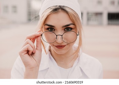 Young female nurse standing outside hospital infirmary. Gorgeous doctor woman dressed white medical gown and cap wear eyeglasses posing, smiling, looking at the camera.
