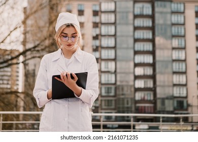Young female nurse standing outside hospital infirmary writing patient illness history in journal. Doctor woman dressed white medical gown and cap wear eyeglasses posing, fill patient paper document
