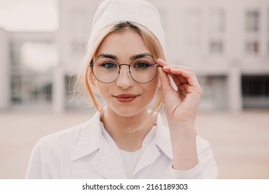 Young female nurse standing outside hospital infirmary. Gorgeous doctor woman dressed white medical gown and cap wear eyeglasses posing, smiling, looking at the camera.