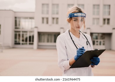 Young female nurse standing outside hospital infirmary. Gorgeous doctor woman dressed white medical gown and face shield, stethescope on neck, writing patient illness history in journal