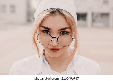 Young female nurse standing outside hospital infirmary. Gorgeous doctor woman dressed white medical gown and cap wear eyeglasses posing, smiling, looking at the camera.
