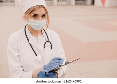 Young female nurse standing outside hospital infirmary writing patient illness history in journal. Doctor woman dressed white medical gown, rubber blue gloves, stethescope and face mask fill paper doc