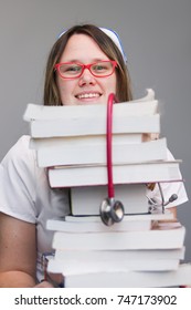 Young Female Nurse Student Portrait Wearing White Scrubs, Cap Holding Tall Stack Of  Nursing Textbooks 