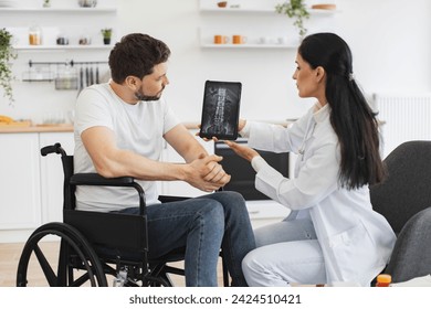 Young female nurse talking to mature man wheelchair user, showing x-ray scan of spine on modern tablet. Adult Caucasian woman doctor checking up condition of mature bearded male patient at home. - Powered by Shutterstock