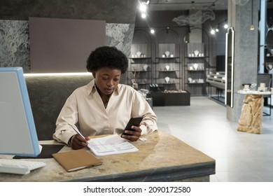 Young Female Owner Of Jewelry Store Or Boutique With Smartphone Working With Papers In Front Of Computer Monitor