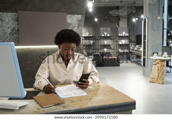 Young female\
owner of jewelry store or boutique with smartphone working with\
papers in front of computer\
monitor