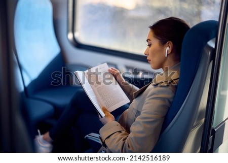 Young female passenger listening music on earbuds and reading book while traveling by train.