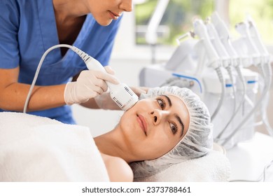 Young female patient experiencing facial cryotherapy procedure using ice hammer to tighten skin in aesthetic medicine clinic. Advanced hardware cosmetology techniques