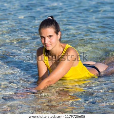 Young female person lying in clear transparent sea water in morning time. Girl in yellow tank top lies on shallow and looks at camera