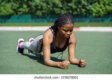 Young Female Person In Sportswear Does Plank Exercise To Strengthen Body On Green Turf Flooring. African American Woman With Long Braids Enjoys Training On Sports Ground Near City Park Closeup