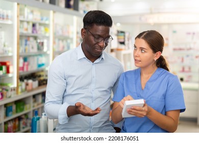 Young Female Pharmacist Advises A Man Visitor A Good Skin Care Remedy, Standing In The Sales Hall Of A Pharmacy