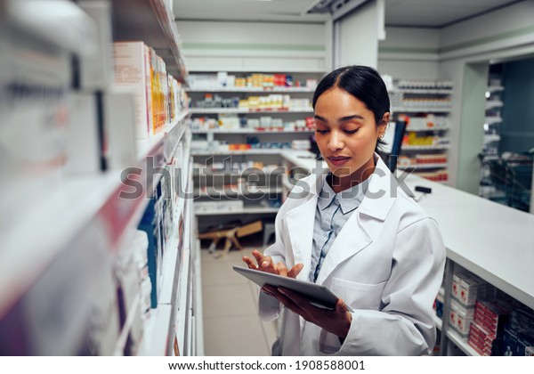 Young female pharmacist checking inventory of\
medicines in pharmacy using digital tablet wearing labcoat standing\
behind counter