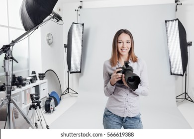 Young female photographer posing in the photo studio, she is smiling and holding a professional digital camera