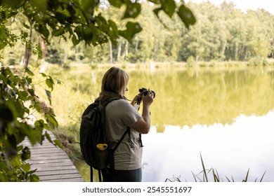 A young female photographer uses a camera to capture a serene lake scene. Travel and hobby concept. Woman with backpack is photographing a landscape near to river - Powered by Shutterstock