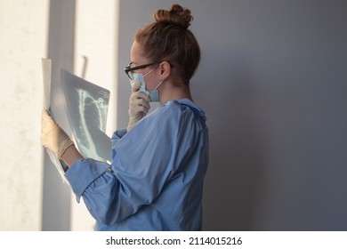 Young Female Physician Looking At Xray Of Lungs And Thinking. Doctor In Mask And Gloves Doing Cancer Or Covid Diagnostics. Tuberculosis Or Pneumonia Conditions. Scan Of Respiratory Organs.