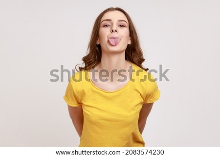 Young female with pleasant appearance wearing yellow T-shirt looking at camera with playful childish facial expression and showing tongue out. Indoor studio shot isolated on gray background.