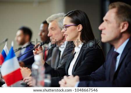 Young female politician in formalwear talking in microphone while making report at political conference