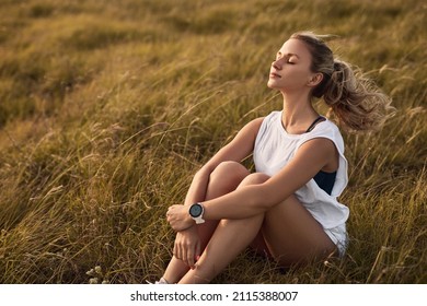 Junge Frau mit Pferdschwanz, die Knie umhüllt und mit geschlossenen Augen frischer Wind genießt, während sie auf trockenem Gras auf dem Land sitzend