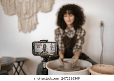 Young female potter sculpting a jug using a potter's wheel while recording a video blog with a mobile phone. Creative art process and vlogging. - Powered by Shutterstock