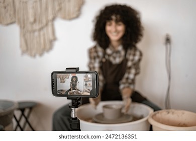 Young female potter sculpts pottery on wheel while recording video blog. Creative process of handmade ceramics captured on smartphone - Powered by Shutterstock