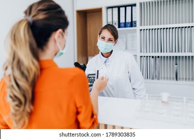 Young female practitioner or nurse with face protective mask working at clinic reception desk. She is helping to patient to make contactless payment for medical services.