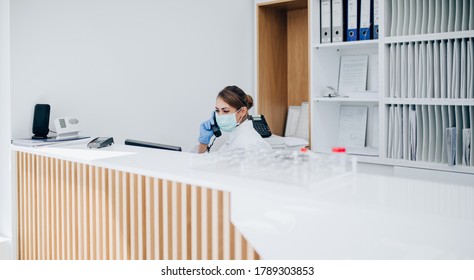 Young Female Practitioner Or Nurse With Face Protective Mask Working At Clinic Reception Desk And Receiving Phone Calls.