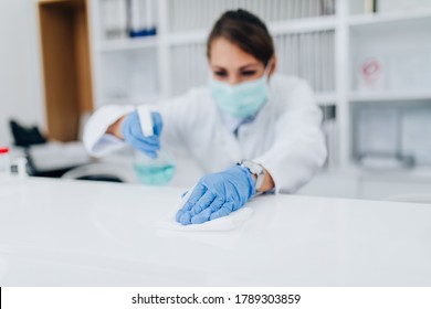 Young Female Practitioner Or Nurse With Face Protective Mask Working At Clinic Reception Desk. She Is Holding Disinfectant Bottle And Cleaining. Selective Focus On Hand.