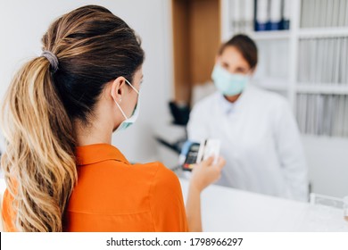 Young Female Practitioner Or Nurse With Face Protective Mask Working At Clinic Reception Desk. She Is Helping To Patient To Make Contactless Payment For Medical Services.