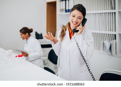 Young female practitioners or nurses with face protective masks working together at clinic reception desk. One nurse answering to the phone calls while another sitting in background.  - Powered by Shutterstock