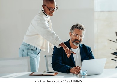 Young female professional assists a seated Caucasian male colleague with work on a laptop in a bright office setting, reflecting teamwork and cooperation in a diverse work environment. - Powered by Shutterstock