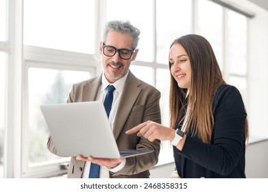 Young female professional points to screen of a laptop while a senior businessman with a jovial expression looks on, indicative of a collaborative and educational moment in a light-filled office space - Powered by Shutterstock