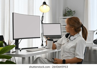 A young female programmer takes a coffee break while working at a clean dual-screen workstation.