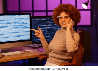 Young female programmer talking by phone and pointing at computer screen in office at night