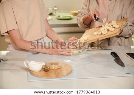 Young female putting banana slices into glass bowl before mixing them with other icecream ingredients during home cooking masterclass