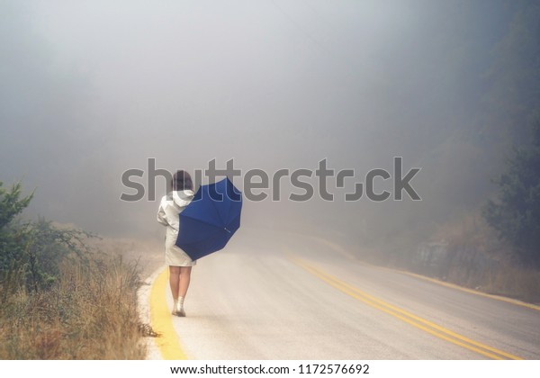 Young female in a\
raincoat on the road in the fog. Travel of women in the raincoat\
hitchhiking in the rain