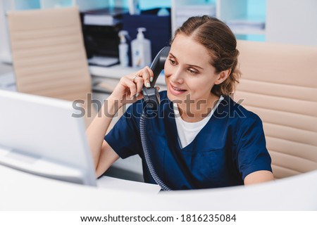 Young female receptionist talking on phone in clinic while sitting and looking on pc monitor