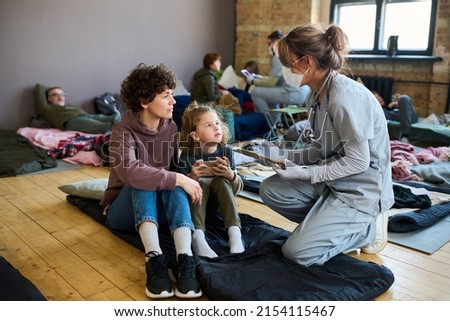 Young female refugee and her little son consulting with healthcare worker in uniform, gloves and protective mask while sitting on sleeping place Foto stock © 