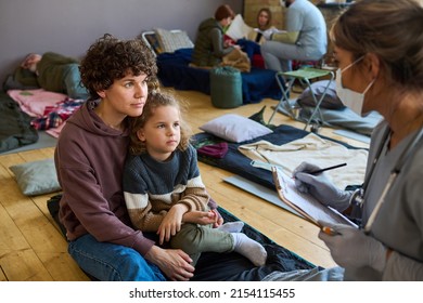 Young Female Refugee With Little Son Looking At Clinician In Uniform And Mask Making Notes In Medical Document And Consulting Them