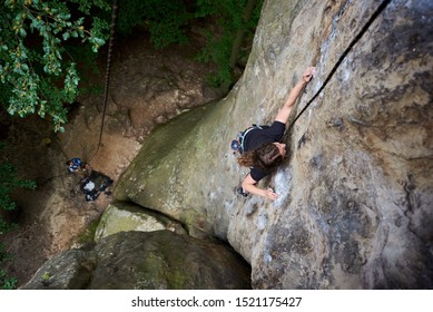 Young female rock climber climbing on cliff with safety harness and rope. Woman overcoming difficulties on rocks. Extreme summer outdoors on difficult climbing route on rock. Sport in nature. Top view