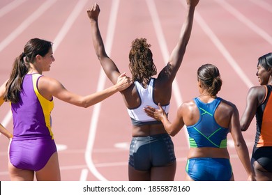 Young Female Runner Cheering Her Victory With Her Arms Raised Overhead While Her Competitors Congratulate Her At The Race Track