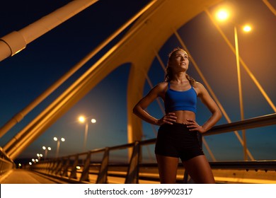 Young Female Runner Confidently Standing With Hands On Hips On Illuminated Bridge At Night And Looking Upwards. Athletic Woman Getting Ready To Exercise To Maintain Shape, Weight And Healthful Mind